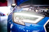 Close-up view of a blue car's headlight with the hood open, revealing engine components. The headlight is brightly illuminated, and the surrounding area seems to be a garage or workshop setting, with blurred background elements.