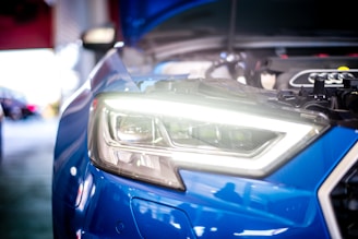 Close-up view of a blue car's headlight with the hood open, revealing engine components. The headlight is brightly illuminated, and the surrounding area seems to be a garage or workshop setting, with blurred background elements.