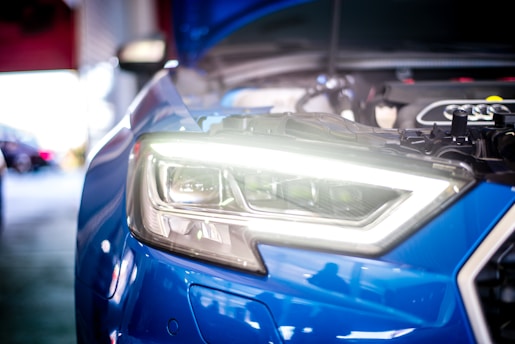 Close-up view of a blue car's headlight with the hood open, revealing engine components. The headlight is brightly illuminated, and the surrounding area seems to be a garage or workshop setting, with blurred background elements.