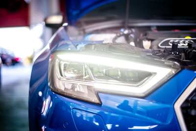 Close-up view of a blue car's headlight with the hood open, revealing engine components. The headlight is brightly illuminated, and the surrounding area seems to be a garage or workshop setting, with blurred background elements.