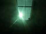 Bright sunlight streaming through a set of horizontal blinds, casting striped shadows on the surrounding surfaces. The light creates a dramatic contrast with the darker areas, and there is a soft, greenish tint in the atmosphere.