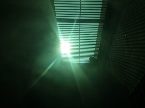 Bright sunlight streaming through a set of horizontal blinds, casting striped shadows on the surrounding surfaces. The light creates a dramatic contrast with the darker areas, and there is a soft, greenish tint in the atmosphere.