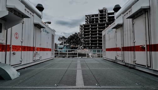 Two large industrial containers with a red stripe are positioned on either side of a metallic walkway. In the background, a partially constructed or dilapidated building stands under a cloudy sky. A lone tree is visible near the center, adding a touch of nature to the industrial setting.