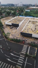 An aerial view of a parking area with several motorcycles parked in designated spots. The scene includes multiple parking lanes outlined in yellow and bordered by concrete barriers. Surrounding the parking space is a road with visible pedestrian crossings and road markings. In the distance, there are various buildings and a green landscape.