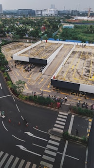 An aerial view of a parking area with several motorcycles parked in designated spots. The scene includes multiple parking lanes outlined in yellow and bordered by concrete barriers. Surrounding the parking space is a road with visible pedestrian crossings and road markings. In the distance, there are various buildings and a green landscape.