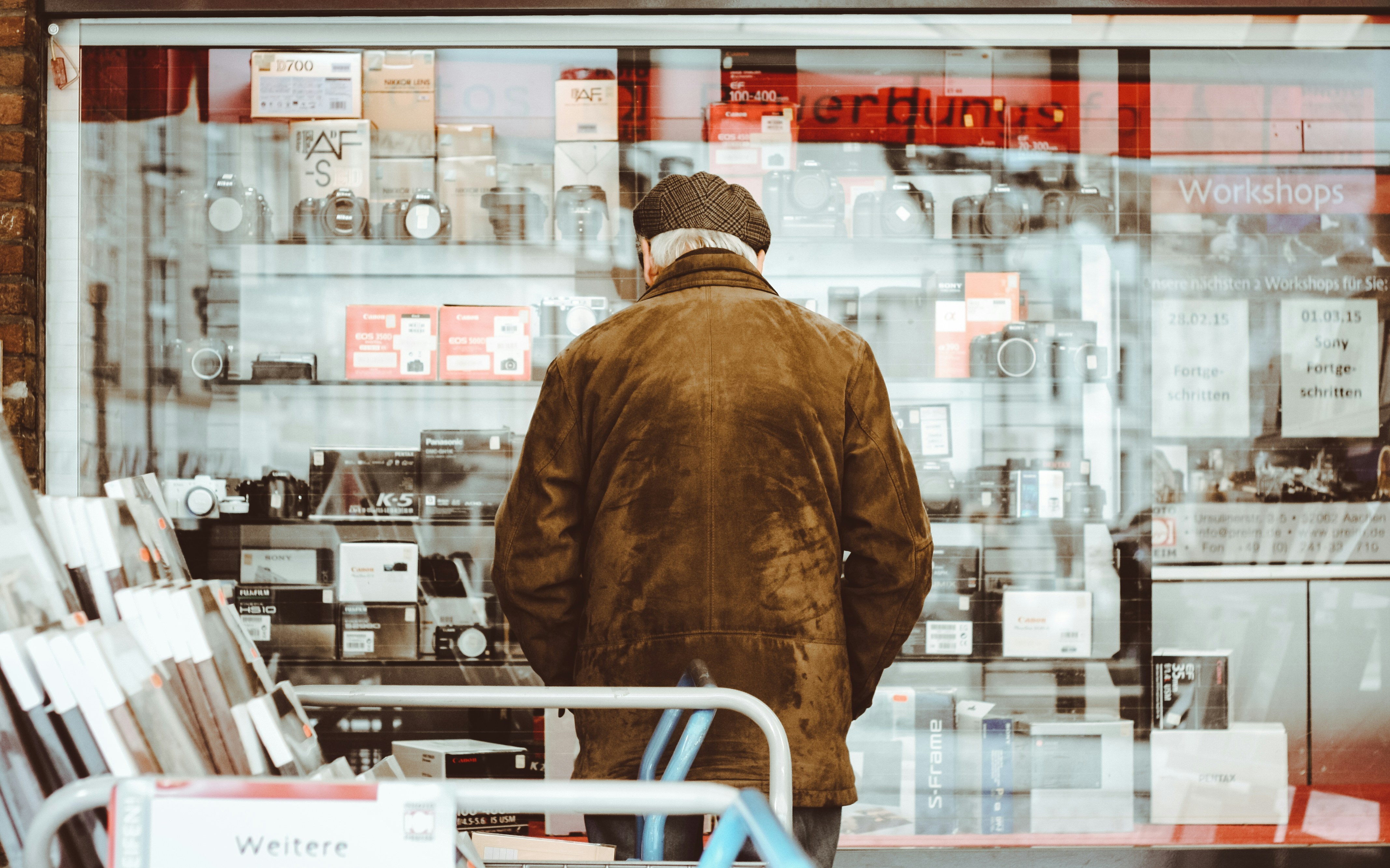 man wearing brown jacket facing clear glass display rack, Old man looking at some used retro cams, ready to be used again.