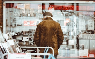 An elderly person wearing a brown coat and hat stands facing a shop window filled with various electronics, including cameras and boxes. The clear glass reflects some light, and a red sign can be seen at the top inside the store. The shelves are neatly arranged with multiple items placed on them, while advertisements and notices are visible on the glass.