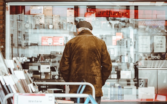 An elderly person wearing a brown coat and hat stands facing a shop window filled with various electronics, including cameras and boxes. The clear glass reflects some light, and a red sign can be seen at the top inside the store. The shelves are neatly arranged with multiple items placed on them, while advertisements and notices are visible on the glass.