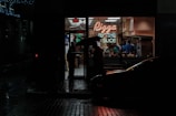A dimly lit street corner at night featuring a small pizza shop with a neon sign that reads 'Pizza by the Slice.' The interior of the shop is bright and contrasts sharply with the dark, wet street outside. A person holding an umbrella stands at the doorway, while several people are visible inside preparing or buying food.
