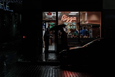 A dimly lit street corner at night featuring a small pizza shop with a neon sign that reads 'Pizza by the Slice.' The interior of the shop is bright and contrasts sharply with the dark, wet street outside. A person holding an umbrella stands at the doorway, while several people are visible inside preparing or buying food.