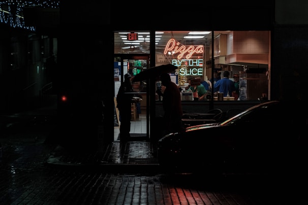 A dimly lit street corner at night featuring a small pizza shop with a neon sign that reads 'Pizza by the Slice.' The interior of the shop is bright and contrasts sharply with the dark, wet street outside. A person holding an umbrella stands at the doorway, while several people are visible inside preparing or buying food.