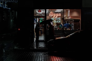 A dimly lit street corner at night featuring a small pizza shop with a neon sign that reads 'Pizza by the Slice.' The interior of the shop is bright and contrasts sharply with the dark, wet street outside. A person holding an umbrella stands at the doorway, while several people are visible inside preparing or buying food.