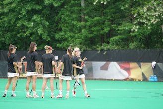 A group of women in black sports jerseys holds lacrosse sticks on a field with green turf. They appear to be in conversation or strategizing before a game or practice session. The background features lush green trees and a black fence with a colorful banner. A cluster of sports balls is visible near the fence, and there are blue water coolers on the right.