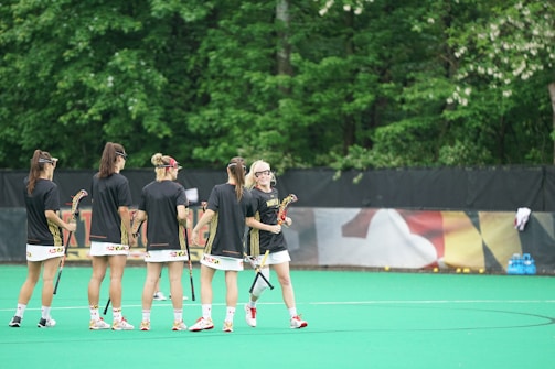A group of women in black sports jerseys holds lacrosse sticks on a field with green turf. They appear to be in conversation or strategizing before a game or practice session. The background features lush green trees and a black fence with a colorful banner. A cluster of sports balls is visible near the fence, and there are blue water coolers on the right.