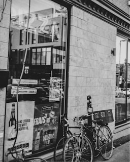 A black and white photograph of a storefront window displaying various advertisements. Two bicycles are parked in front of the window, one of which has a basket attached. The reflection of surrounding buildings can be seen in the glass.