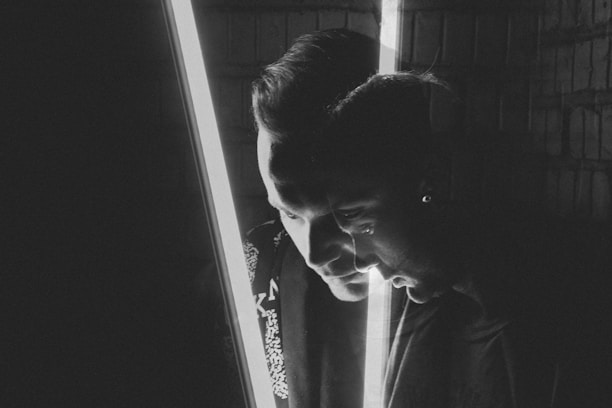A grayscale portrait features a double exposure effect, capturing two profiles of a person facing opposite directions. A bright vertical light bisects the image, adding dramatic contrast.