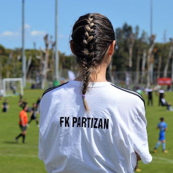 A person with braided hair is wearing a white T-shirt with 'FK Partizan' printed on the back, observing a soccer game. The scene takes place on a grass field with blurred figures of players and a referee in the background. The day is sunny with a blue sky and tall poles are visible.