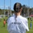 A person with braided hair is wearing a white T-shirt with 'FK Partizan' printed on the back, observing a soccer game. The scene takes place on a grass field with blurred figures of players and a referee in the background. The day is sunny with a blue sky and tall poles are visible.
