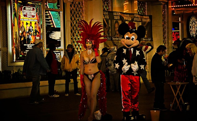 A street scene at night with bright lights and people in costume. A performer in a red feathered headdress and a revealing outfit stands next to a person in a Mickey Mouse costume. Several passersby are visible, and there are illuminated signs and posters in the background.