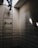 A person is climbing a metal ladder that leads to an opening in the ceiling. The surrounding area is an enclosed space with bare concrete brick walls. Light is entering through the opening, casting sharp, angled shadows on the walls.