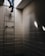 A person is climbing a metal ladder that leads to an opening in the ceiling. The surrounding area is an enclosed space with bare concrete brick walls. Light is entering through the opening, casting sharp, angled shadows on the walls.