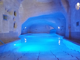 A serene indoor swimming pool with a cave-like ceiling and walls, creating a tranquil and mysterious atmosphere. The pool is illuminated with blue lighting, enhancing the calm ambiance. The architecture has a unique, natural stone appearance, with small recessed lights on the sides.