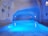 A serene indoor swimming pool with a cave-like ceiling and walls, creating a tranquil and mysterious atmosphere. The pool is illuminated with blue lighting, enhancing the calm ambiance. The architecture has a unique, natural stone appearance, with small recessed lights on the sides.