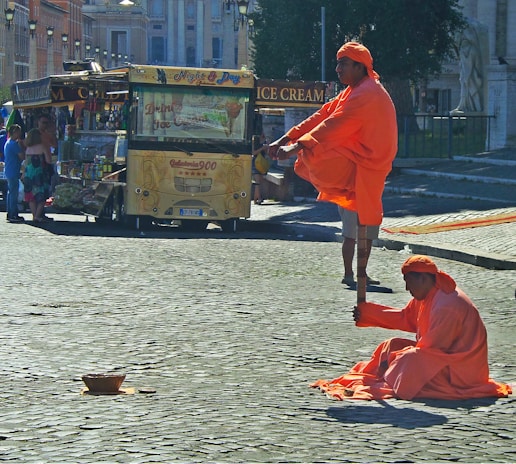 Two individuals dressed in orange robes are performing a street act involving levitation on a cobblestone street. One person sits on the ground holding a pole, while the other appears to be elevated above the ground with legs crossed, supported by the pole. An ice cream and food stall is visible in the background, with people gathered around it. The setting appears to be a public urban area during daylight.