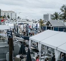A bustling marina filled with large yachts and numerous tents, showcasing a vibrant nautical event. People are milling about, some observing the boats while others are inside the tents. Palm trees are interspersed throughout, and buildings are visible in the background.