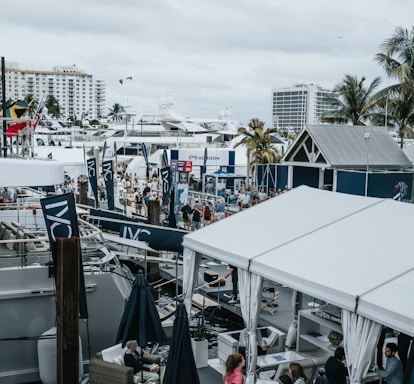 A bustling marina filled with large yachts and numerous tents, showcasing a vibrant nautical event. People are milling about, some observing the boats while others are inside the tents. Palm trees are interspersed throughout, and buildings are visible in the background.