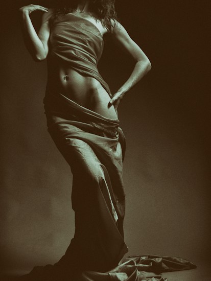A sepia-toned photograph featuring a partially draped figure. The lighting softly highlights the curves and contours of the body, and the flowing fabric adds a sense of movement.