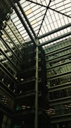A modern architectural interior featuring a large atrium with a glass ceiling. The structure includes multiple floors of offices with glass windows. Some green, plant-covered walls add a touch of nature amidst the industrial design.