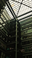A modern architectural interior featuring a large atrium with a glass ceiling. The structure includes multiple floors of offices with glass windows. Some green, plant-covered walls add a touch of nature amidst the industrial design.