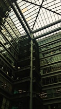 A modern architectural interior featuring a large atrium with a glass ceiling. The structure includes multiple floors of offices with glass windows. Some green, plant-covered walls add a touch of nature amidst the industrial design.