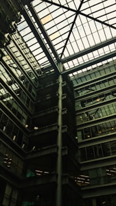 A modern architectural interior featuring a large atrium with a glass ceiling. The structure includes multiple floors of offices with glass windows. Some green, plant-covered walls add a touch of nature amidst the industrial design.