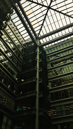 A modern architectural interior featuring a large atrium with a glass ceiling. The structure includes multiple floors of offices with glass windows. Some green, plant-covered walls add a touch of nature amidst the industrial design.