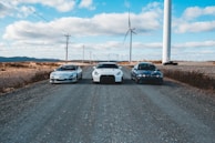 Three cars are parked on a gravel road under a blue sky with scattered clouds. Wind turbines are standing tall in the background, lined across a barren landscape. The scene conveys a mix of technology and nature with the use of wind energy highlighted by the turbines.