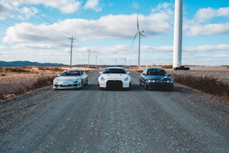 Three cars are parked on a gravel road under a blue sky with scattered clouds. Wind turbines are standing tall in the background, lined across a barren landscape. The scene conveys a mix of technology and nature with the use of wind energy highlighted by the turbines.