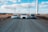 Three cars are parked on a gravel road under a blue sky with scattered clouds. Wind turbines are standing tall in the background, lined across a barren landscape. The scene conveys a mix of technology and nature with the use of wind energy highlighted by the turbines.
