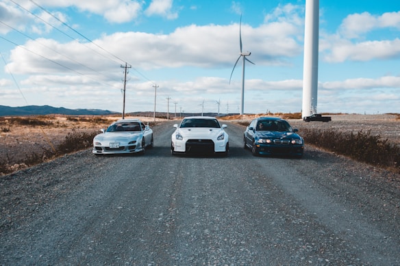Three cars are parked on a gravel road under a blue sky with scattered clouds. Wind turbines are standing tall in the background, lined across a barren landscape. The scene conveys a mix of technology and nature with the use of wind energy highlighted by the turbines.