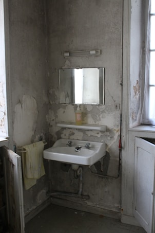 A small, neglected bathroom corner with peeling walls. The scene features a white porcelain sink mounted to the wall, with visible plumbing beneath. Above the sink, a simple rectangular mirror is fixed to the wall, alongside an old fluorescent light fixture. A yellowing towel hangs on a rack, and the overall condition suggests disrepair or abandonment, with cracked plaster and visible dirt.