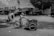 A man pushes a three-wheeled cart on a rough, muddy street in front of various small businesses. The scene appears to be in a developing area, with multiple signs for automotive and industrial services in the background. The buildings are slightly dilapidated and the area has a gritty, industrial feel.