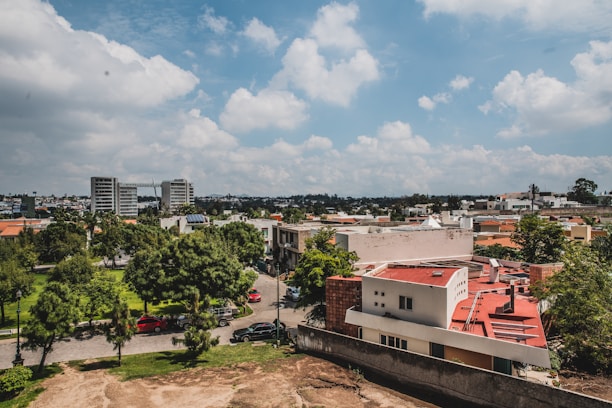 Urban landscape featuring a cluster of buildings with predominantly red and white rooftops. There is greenery with a variety of trees lining the streets and a well-maintained park visible from above. A few parked cars are visible along the roadways. In the background, a city skyline with taller structures can be seen under a partly cloudy blue sky.