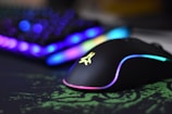 A sleek gaming mouse with RGB lighting effects sits on a green and black mousepad. In the background, a blurred view of a keyboard with vibrant RGB lighting can be seen, adding a futuristic and colorful ambiance.