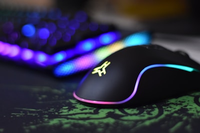 A sleek gaming mouse with RGB lighting effects sits on a green and black mousepad. In the background, a blurred view of a keyboard with vibrant RGB lighting can be seen, adding a futuristic and colorful ambiance.