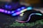 A sleek gaming mouse with RGB lighting effects sits on a green and black mousepad. In the background, a blurred view of a keyboard with vibrant RGB lighting can be seen, adding a futuristic and colorful ambiance.