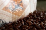 A close-up view of a coffee bag partially visible, surrounded by a scattered pile of roasted coffee beans. The bag has a geometric design with the words 'Boldly Go' and an established date of 2012 printed on it. The colors are warm, with a focus on browns and beiges.