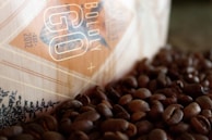 A close-up view of a coffee bag partially visible, surrounded by a scattered pile of roasted coffee beans. The bag has a geometric design with the words 'Boldly Go' and an established date of 2012 printed on it. The colors are warm, with a focus on browns and beiges.