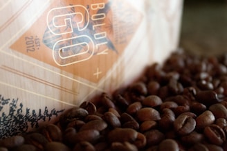 A close-up view of a coffee bag partially visible, surrounded by a scattered pile of roasted coffee beans. The bag has a geometric design with the words 'Boldly Go' and an established date of 2012 printed on it. The colors are warm, with a focus on browns and beiges.