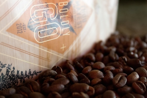 A close-up view of a coffee bag partially visible, surrounded by a scattered pile of roasted coffee beans. The bag has a geometric design with the words 'Boldly Go' and an established date of 2012 printed on it. The colors are warm, with a focus on browns and beiges.